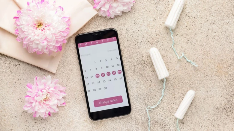 Telefon s kalendářem menstruace vedle tampónů na texturovaném povrchu.