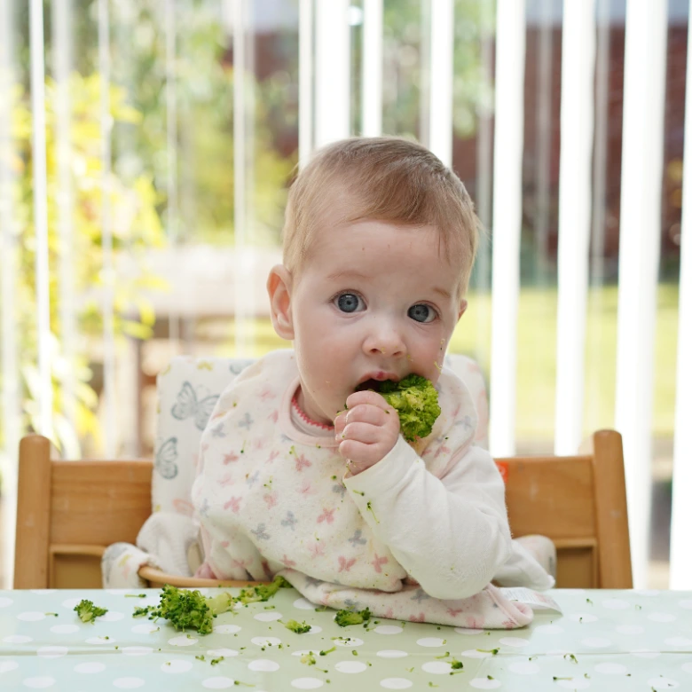 Malý dítě se samostatně krmit brokolicí v jídelní židli.