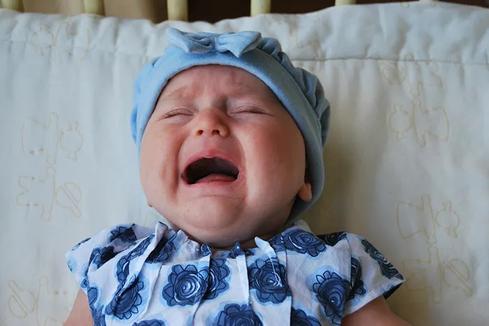 Pláčající dítě v modré čepici na bílém povlečení.