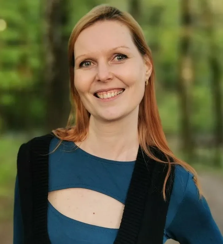 Usměvavá žena v modrém tričku stojí venku před lesním pozadím.