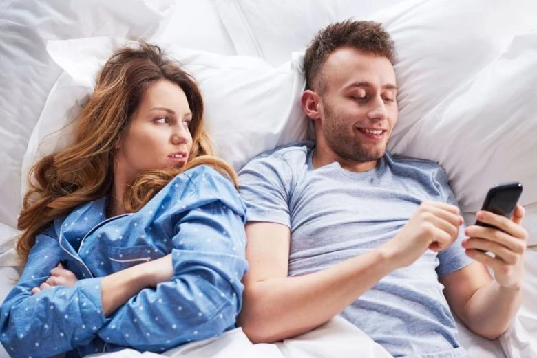 Muž a žena leží v posteli, muž kouká na telefon.