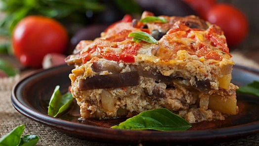 Kousek lasagne s lilkem na talíři ozdobený bazalkou.