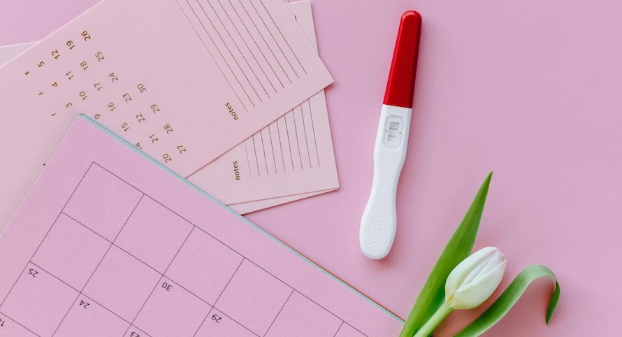 Těhotenský test s kalendářem menstruačního cyklu na růžovém pozadí.