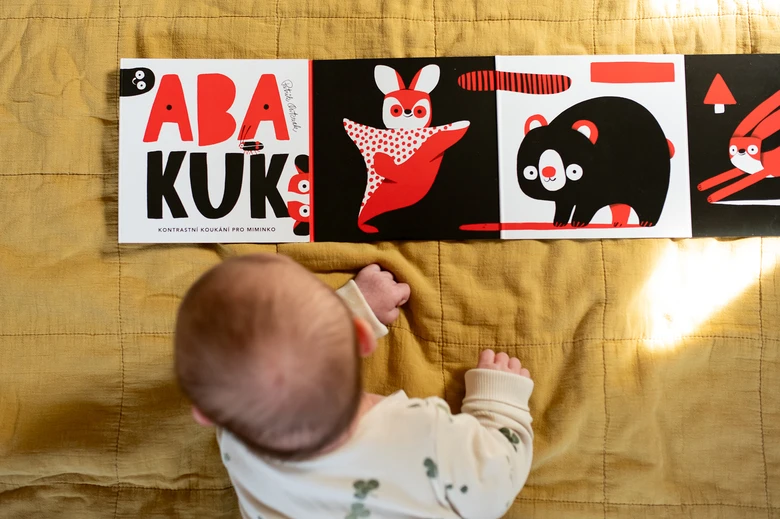 Miminko leží na žlutém prostěradle a dívá se do kontrastní knihy pro děti.