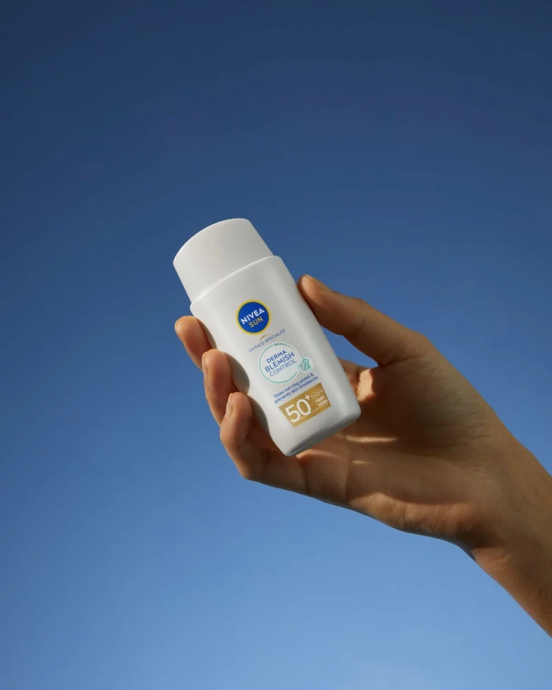 Ruka drží opalovací krém s vysokým SPF proti modré obloze.