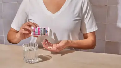 Žena bere probiotika z lahvičky do ruky vedle sklenice vody.