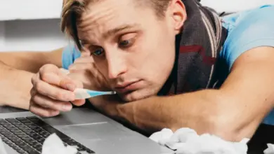 Muž s teploměrem u úst leží na laptopu při nachlazení.