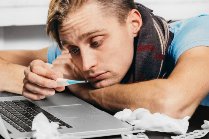 Muž s teploměrem u úst leží na laptopu při nachlazení.