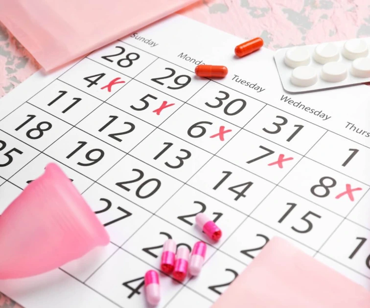 Kalendář s označením menstruace a antikoncepční pilulky na růžovém pozadí.