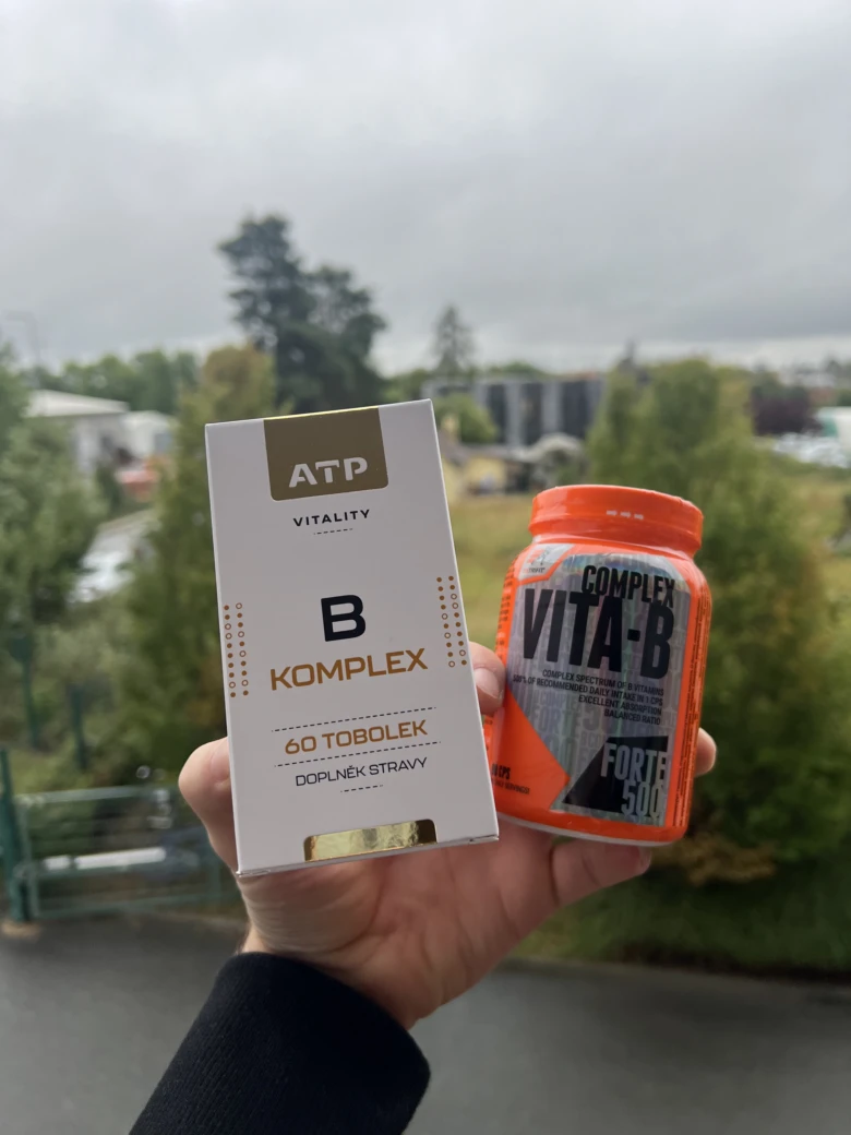 Ruka drží krabičku vitamínu B komplex vedle oranžového sáčku.