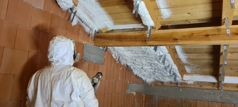 Muž v ochranném obleku stříká izolační pěnu na dřevěné trámy.