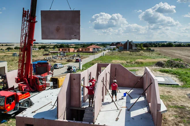 Kran zvedá keramopanel na stavbě hrubé konstrukce s dělníky.