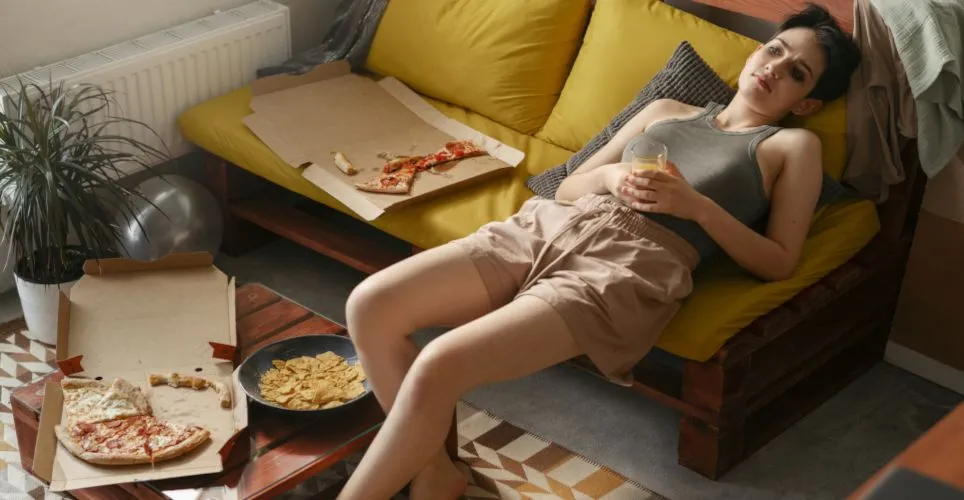 Žena leží na gauči s pizzou a čipy po jídle.